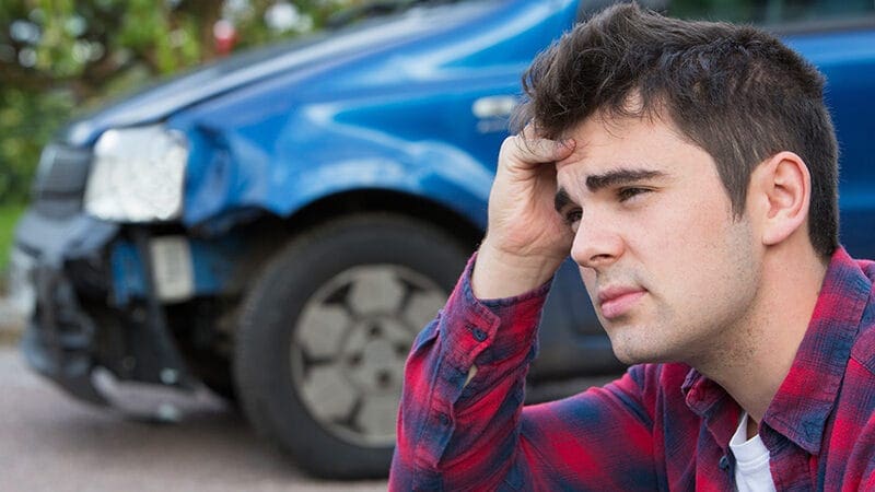 A young man in a red plaid shirt holds his head, looking concerned while sitting near a damaged blue car. The vehicle's front area appears dented, suggesting a recent accident. Greenery is visible in the background.