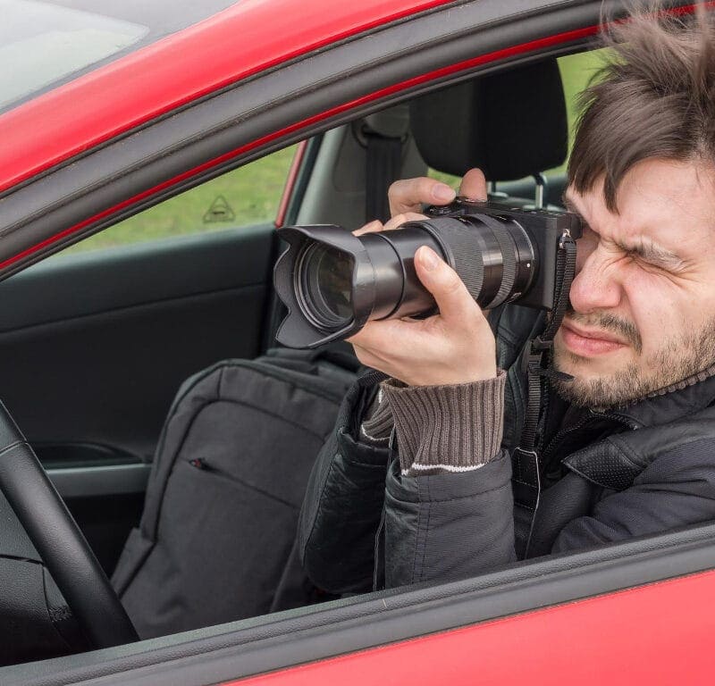 A man sits inside a red car, camera with a large zoom lens in hand, keenly focused on capturing the perfect shot through the open driver's side window. His windswept hair hints at the dedication involved in mastering personal injury surveillance.