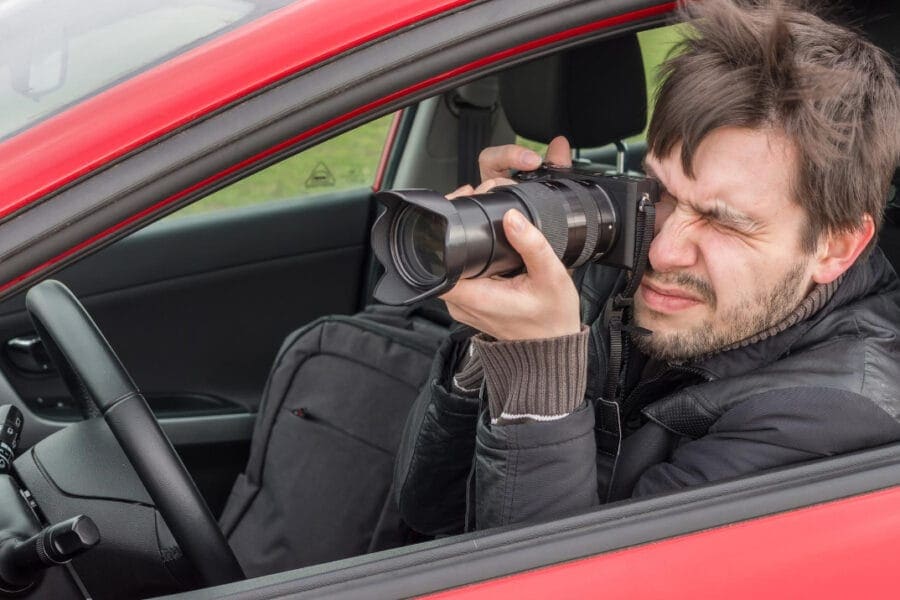 A man sits inside a red car, camera with a large zoom lens in hand, keenly focused on capturing the perfect shot through the open driver's side window. His windswept hair hints at the dedication involved in mastering personal injury surveillance.
