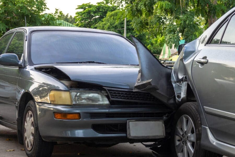Two cars involved in a rear-end collision on a street with greenery in the background. The front car has significant front-end damage, while the rear car has damage to the back. Both vehicles are parked at a slight angle.
