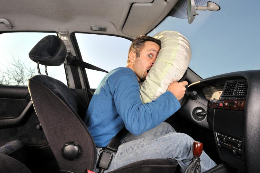 A man in a blue shirt is sitting in the driver's seat of a car, looking surprised as the airbag deploys in front of him. The car's interior is visible, including the steering wheel and dashboard.