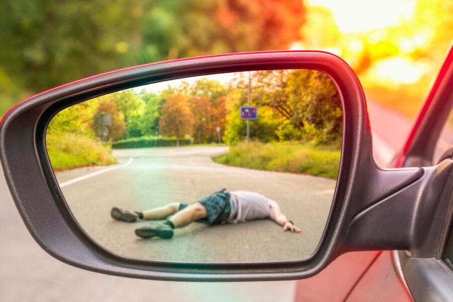 A person is lying on the ground on a curved road, seen through a car's side mirror. Surrounded by greenery and soft sunlight, the scene captures a tense moment with no other visible activity nearby.