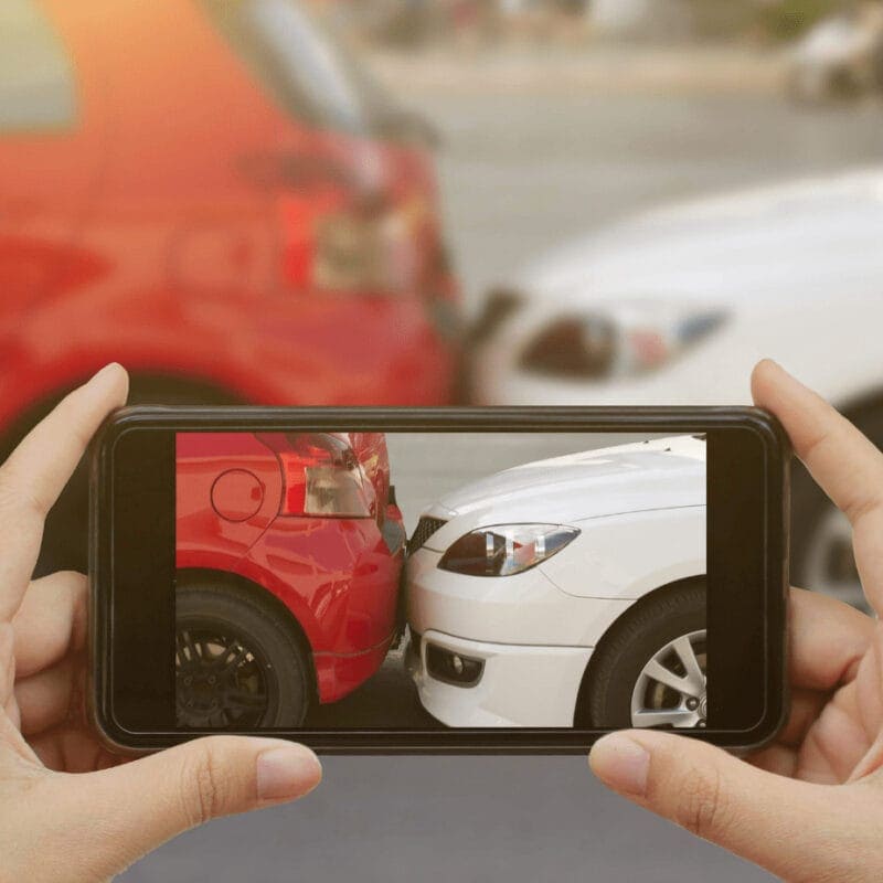 A person holds a smartphone capturing a photo of a minor car accident between a red car and a white car. The cars are bumper to bumper with slight damage visible.