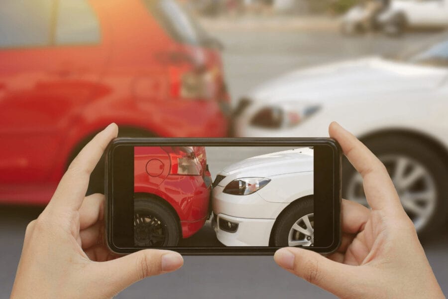 A person holds a smartphone capturing a photo of a minor car accident between a red car and a white car. The cars are bumper to bumper with slight damage visible.