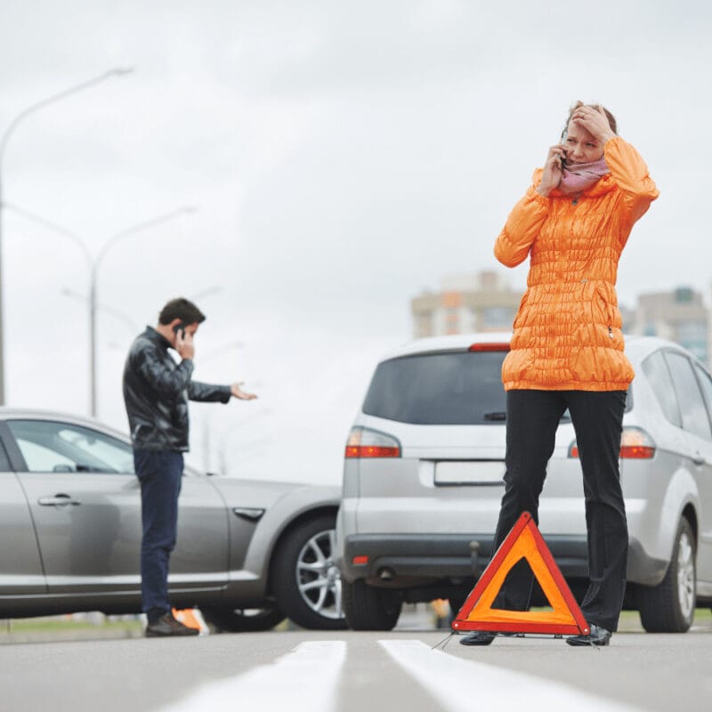 A woman in an orange jacket talks on her phone near a car accident, holding her head. A man stands by another car, gesturing in frustration. A warning triangle is placed on the road. Buildings and streetlights are visible in the background.