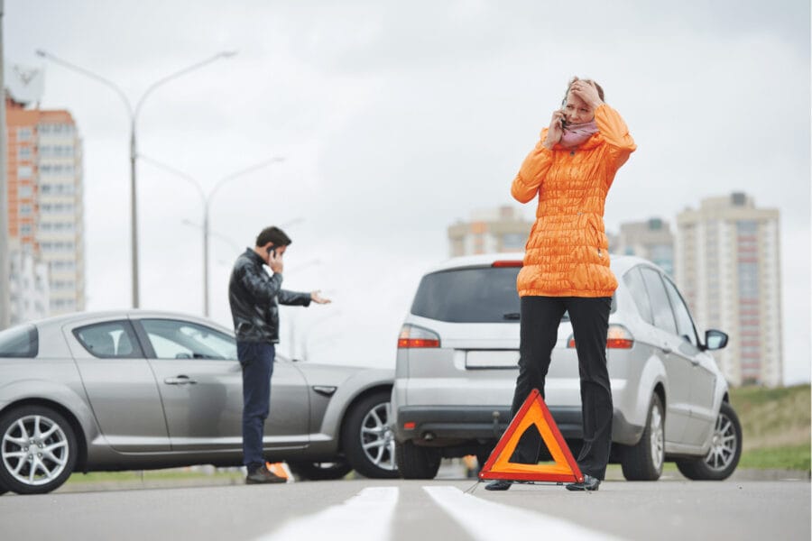 A woman in an orange jacket talks on her phone near a car accident, holding her head. A man stands by another car, gesturing in frustration. A warning triangle is placed on the road. Buildings and streetlights are visible in the background.
