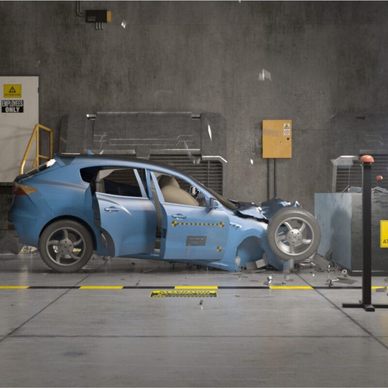 A blue car is crashed into a barrier inside a testing facility. The vehicle's front is heavily damaged, and debris is scattered around. Yellow caution signs and bright lights are positioned nearby, indicating a controlled crash test environment.