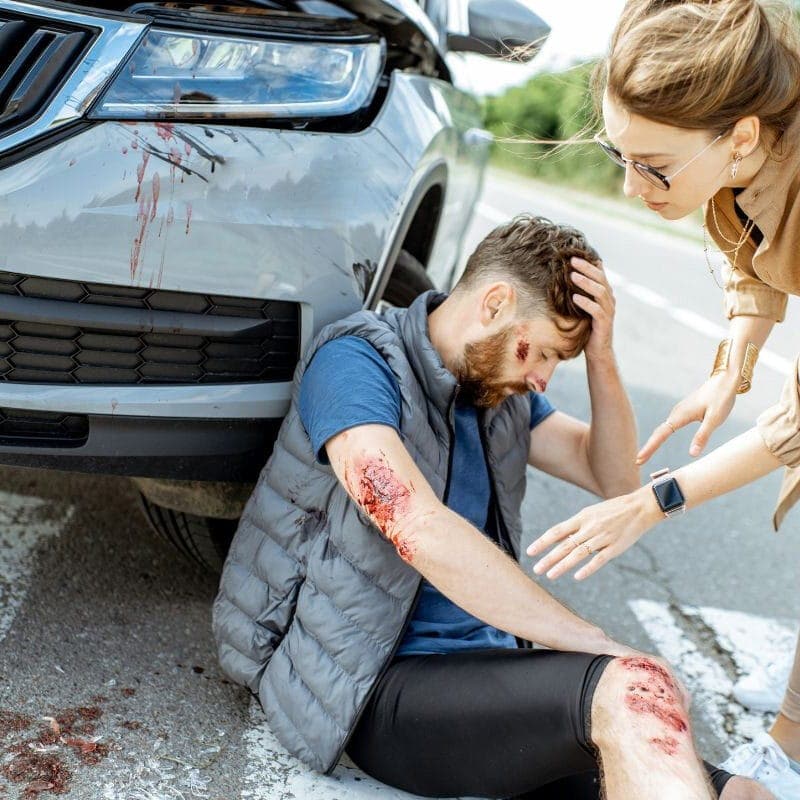 A woman helps an injured man sitting on the ground next to a car with a damaged front. The man has scrapes on his arms and appears dazed. They are in the road on a sunny day, with blood visible on the car.