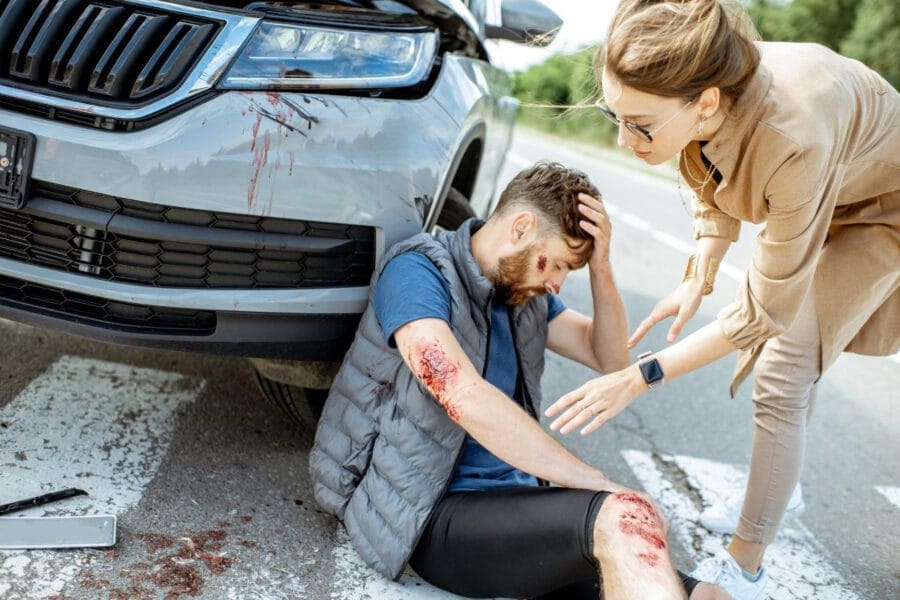 A woman helps an injured man sitting on the ground next to a car with a damaged front. The man has scrapes on his arms and appears dazed. They are in the road on a sunny day, with blood visible on the car.