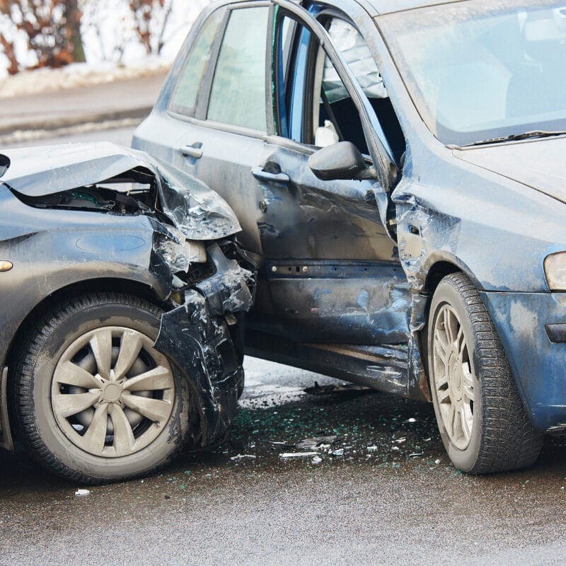 Two cars involved in a collision on a wet road. The front of a black car is heavily damaged, and the side of a blue car is dented. Broken glass is scattered on the ground.