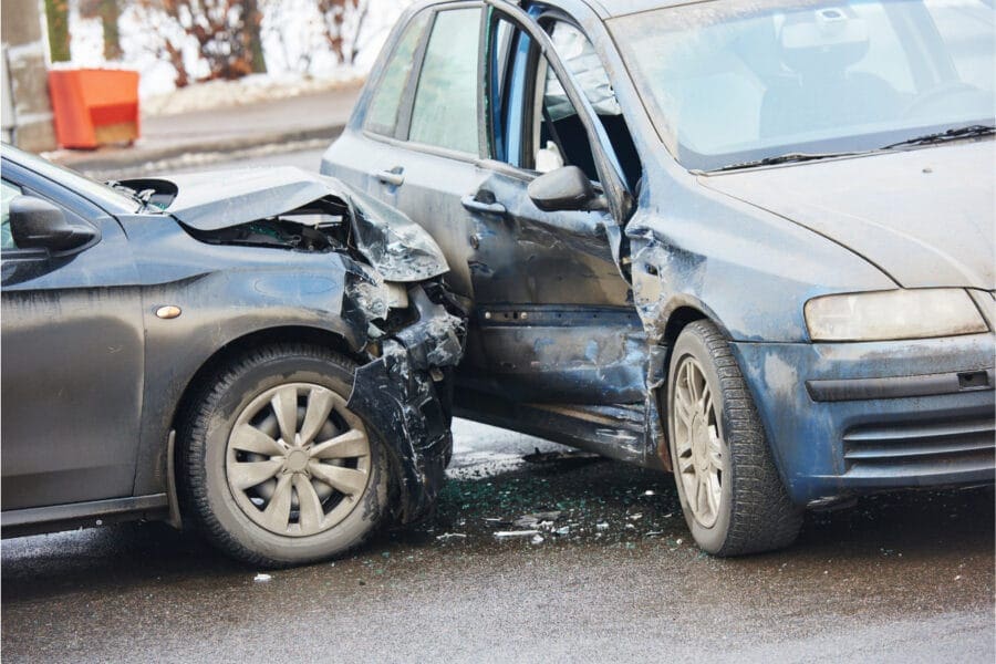 Two cars involved in a collision on a wet road. The front of a black car is heavily damaged, and the side of a blue car is dented. Broken glass is scattered on the ground.