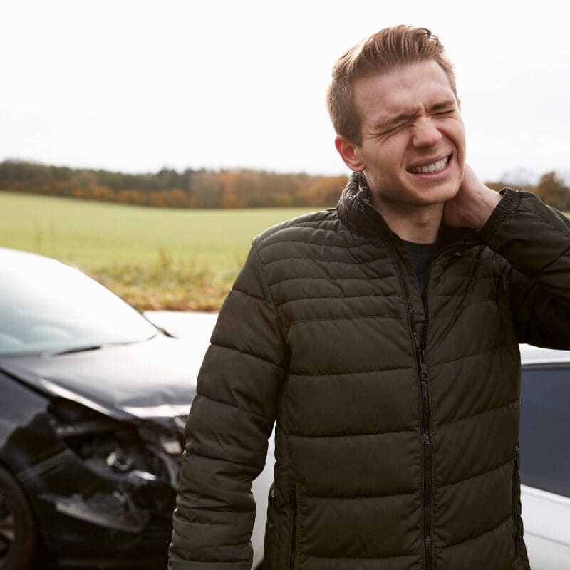 A man stands outside, holding his neck and wincing in pain. In the background, two cars are involved in what is a minor accident on a rural road. The front of the black car is damaged. The countryside setting features trees and fields.