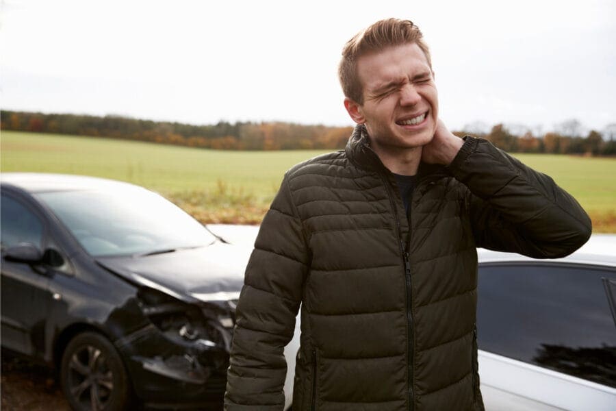 A man stands outside, holding his neck and wincing in pain. In the background, two cars are involved in what is a minor accident on a rural road. The front of the black car is damaged. The countryside setting features trees and fields.