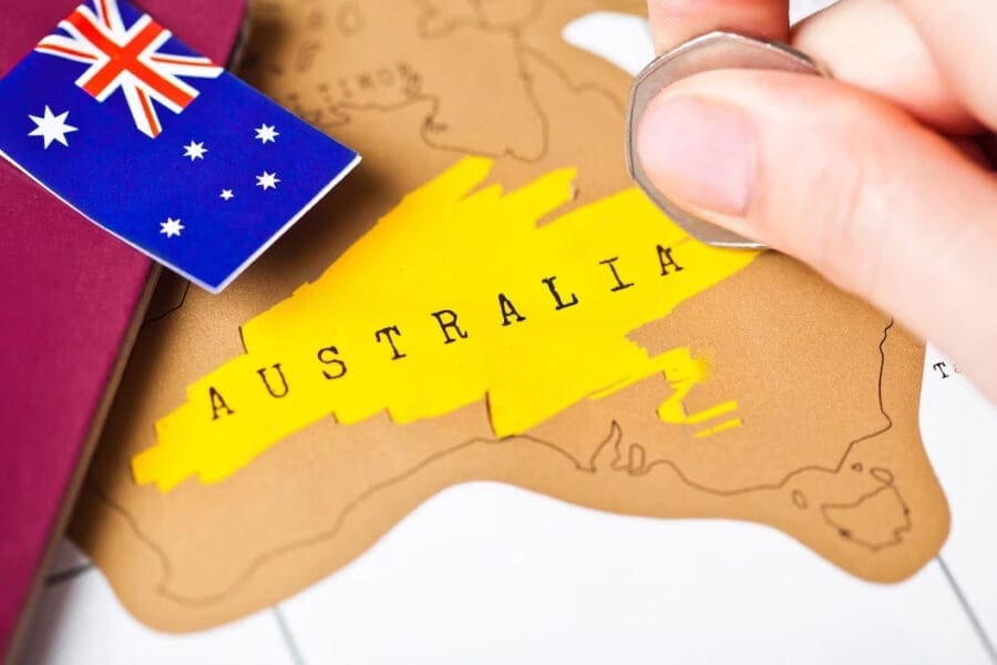 A hand holding a coin scratches off a golden area on a map of Australia, perhaps pondering what state has the most car accidents. Nearby, a small flag card and a maroon travel document suggest upcoming adventures.