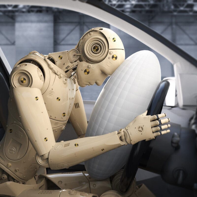 A humanoid robot sits in the driver's seat of a car, its head nestled on an inflated airbag, evaluating what is the best position to survive a car crash. The industrial setting hints at rigorous vehicle safety testing or crash simulation.