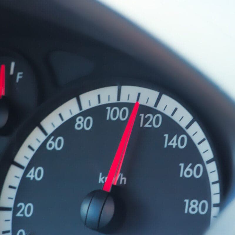 A car dashboard displays a speedometer at 100 km/h, strikingly close to the speed where most car accidents happen. The fuel gauge indicates half a tank. Black dials with white numbers and red needles stand out against the softly blurred background.