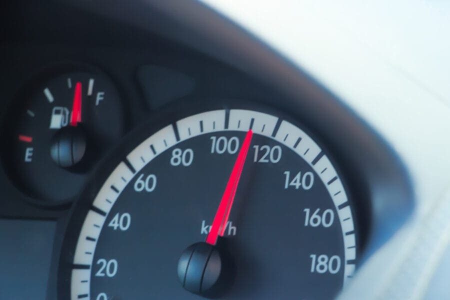 A car dashboard displays a speedometer at 100 km/h, strikingly close to the speed where most car accidents happen. The fuel gauge indicates half a tank. Black dials with white numbers and red needles stand out against the softly blurred background.