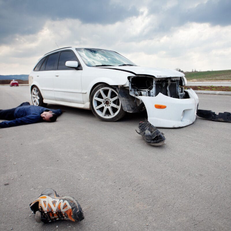 A car accident scene with a damaged white car and a person lying on the ground.
