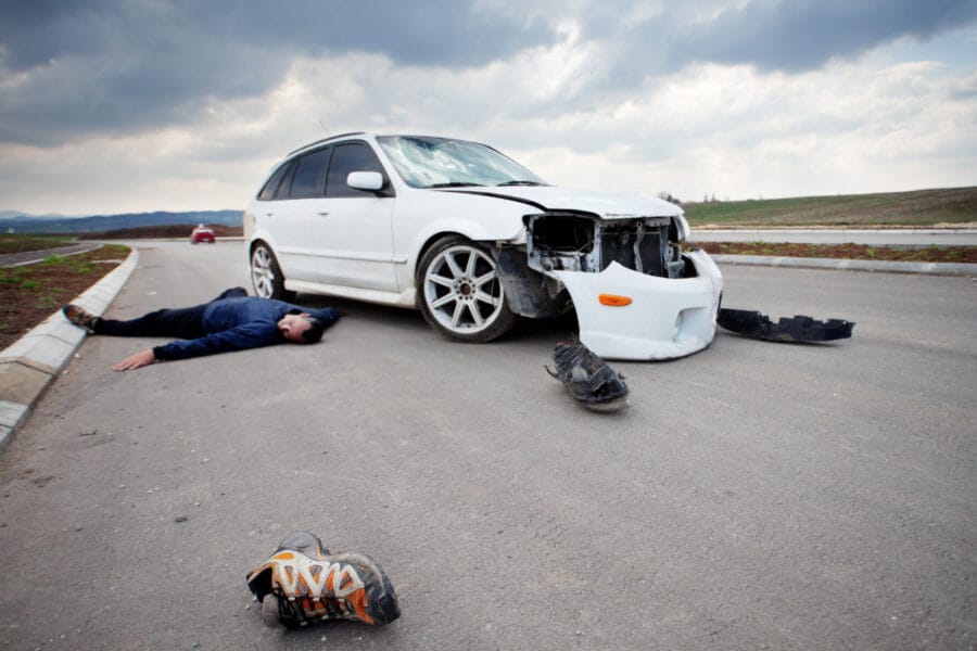 A car accident scene with a damaged white car and a person lying on the ground.