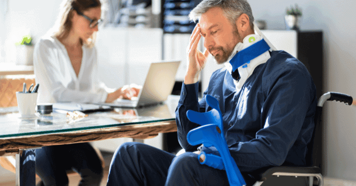 A man with a neck brace and arm support sits in a wheelchair, massaging his temple. A woman in the background is focused on her laptop at a desk, wearing glasses and a light blouse. The scene suggests an office setting.